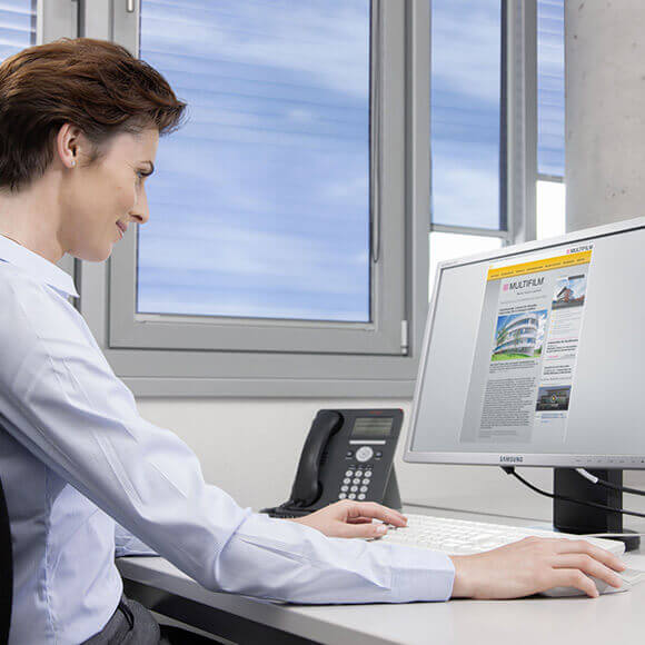 Frau am Computerarbeitsplatz, dahinter Fenster mit Multifilm Rollos